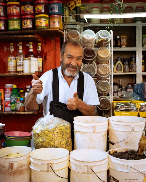 A grocer standing behind a stall of dried goods, giving a thumbs up to the camera.