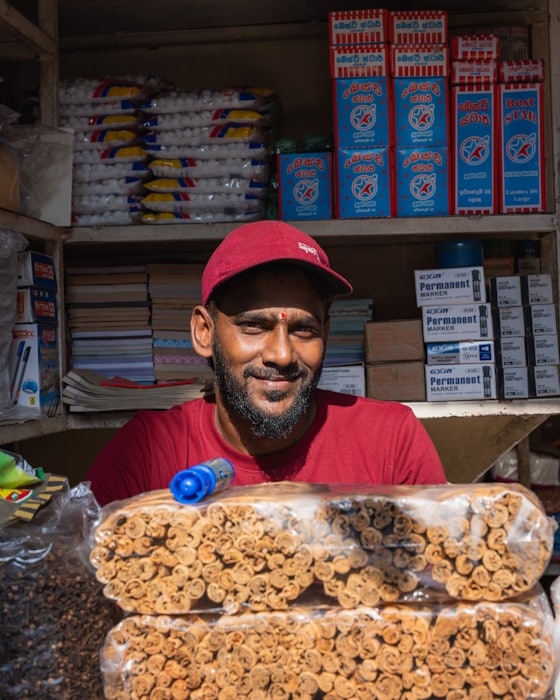 A shopkeeper smiling behind the counter of a small general store
          stocked with packaged goods.